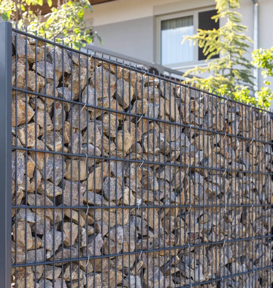 A modern gabion fence constructed from stones creates an elegant boundary. It stands in a contemporary setting, complemented by greenery and a sleek architectural backdrop, showcasing innovation Zaunbau auf Usedom